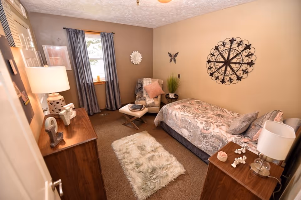 A cozy bedroom with a single bed covered in a patterned comforter and multiple pillows. There is a wooden dresser with a lamp and decorative items on it, and a small wooden nightstand with a lamp and small decor pieces. A comfortable armchair with a pink pillow and a blanket is placed near a window with blue curtains. The walls are beige and decorated with a large ornamental metal wall piece, a butterfly decoration, and a white flower wall art. A fluffy white rug lies on the carpeted floor.