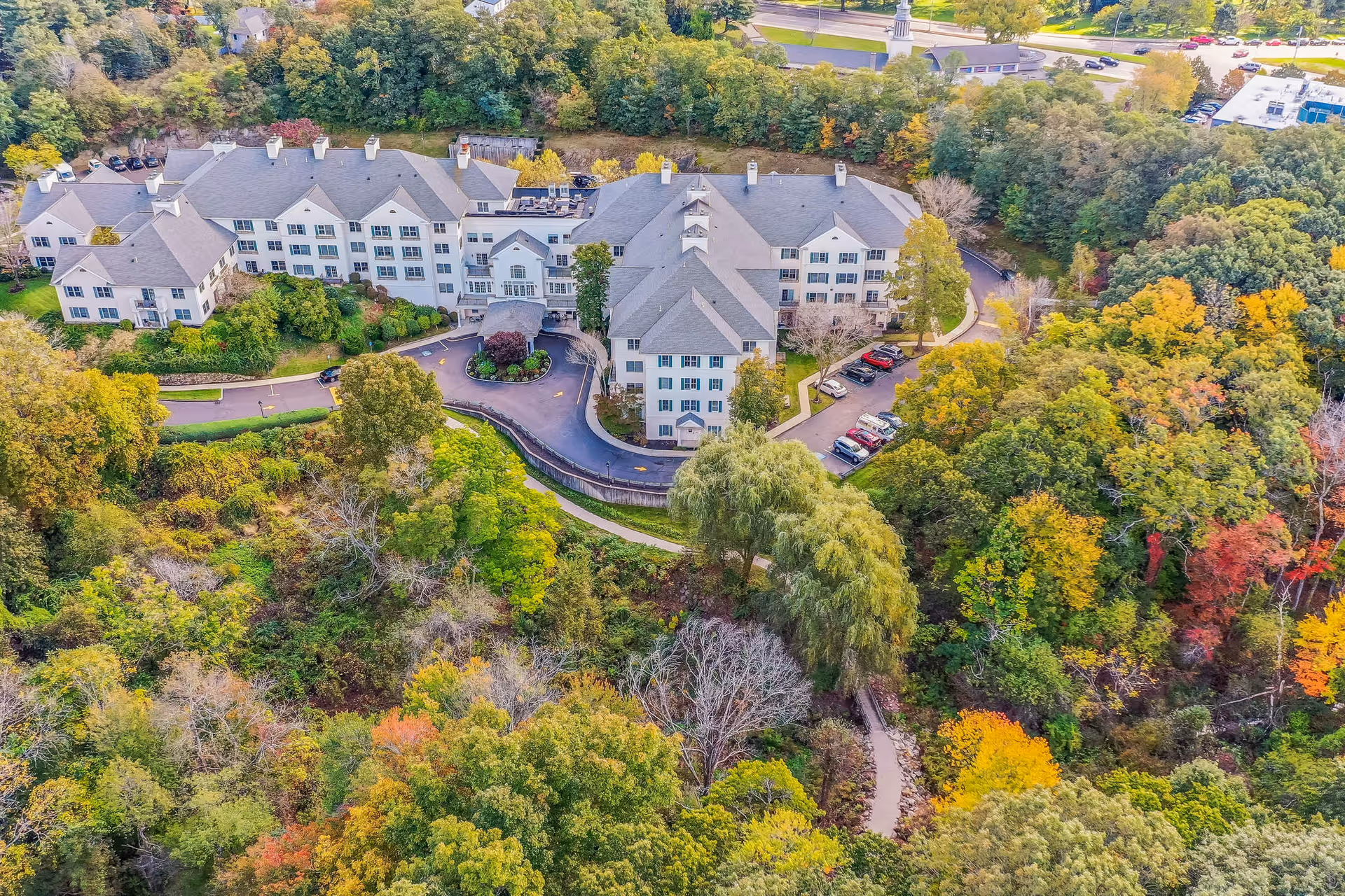 Aerial view of a large senior living facility surrounded by dense trees with autumn foliage. The building has multiple wings with gray roofs and white walls, and there is a circular driveway with parked cars. A walking path winds through the wooded area in front of the building.