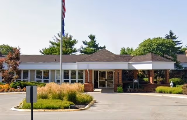 Front exterior view of Cumberland Pointe Health Campus building with a covered entrance, flagpole, and landscaped greenery including bushes and trees under a clear sky.