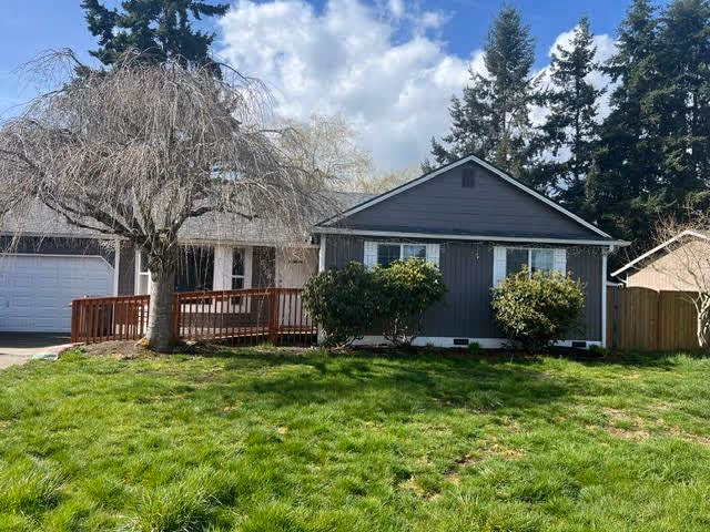 Single-story residential building with a gray exterior, white trim, and a wooden ramp leading to the front door. There are two bushes in front of the building and a leafless tree on the left side. The sky is partly cloudy and there are tall evergreen trees in the background.