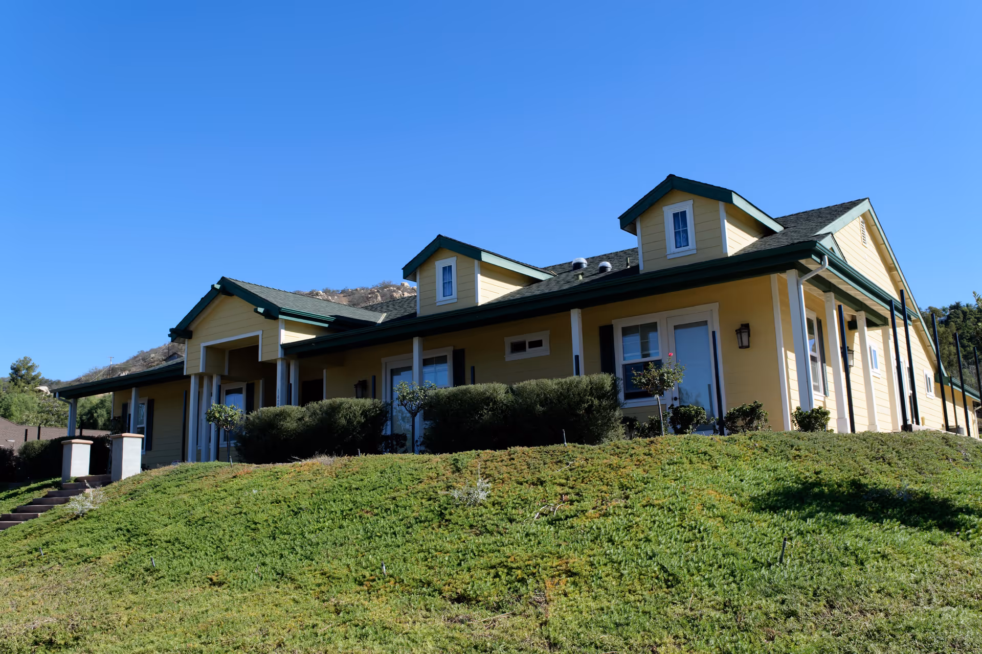 Exterior view of a yellow single-story building with green trim and a dark roof, situated on a grassy hill under a clear blue sky.