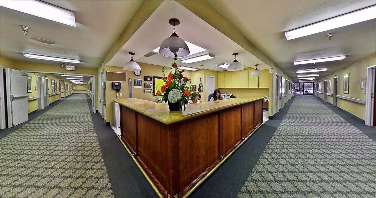 Interior view of a nursing facility hallway with a central wooden reception desk adorned with a flower arrangement. Two staff members are seated behind the desk. The hallway extends on both sides with multiple doors and handrails along the walls. The ceiling has fluorescent lighting and pendant lamps above the desk.