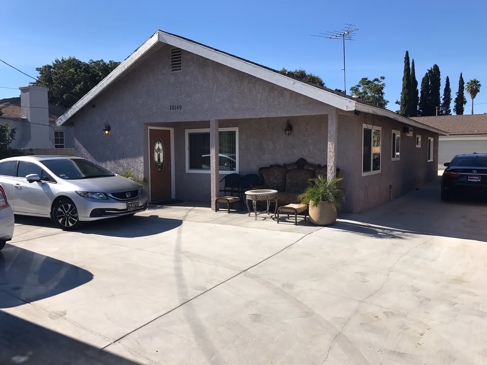 Exterior view of a single-story assisted living facility building with a sloped roof, a front door with a decorative glass panel, a window, and a small seating area with chairs, a table, and a potted plant on a concrete driveway. Two cars are parked in front of the building under a clear blue sky.