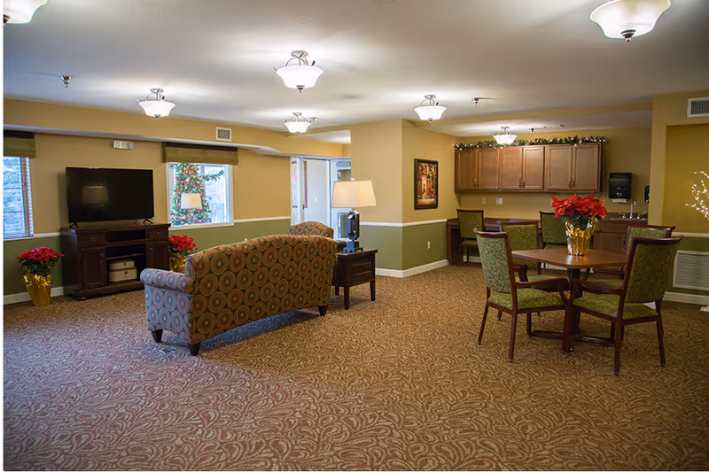 A cozy senior living common area with patterned carpet, a patterned sofa facing a flat-screen TV on a wooden stand, a side table with a lamp, and a round wooden table with four green upholstered chairs. The room has beige and green walls, ceiling lights, windows with blinds, and festive holiday decorations including poinsettia plants and garlands.