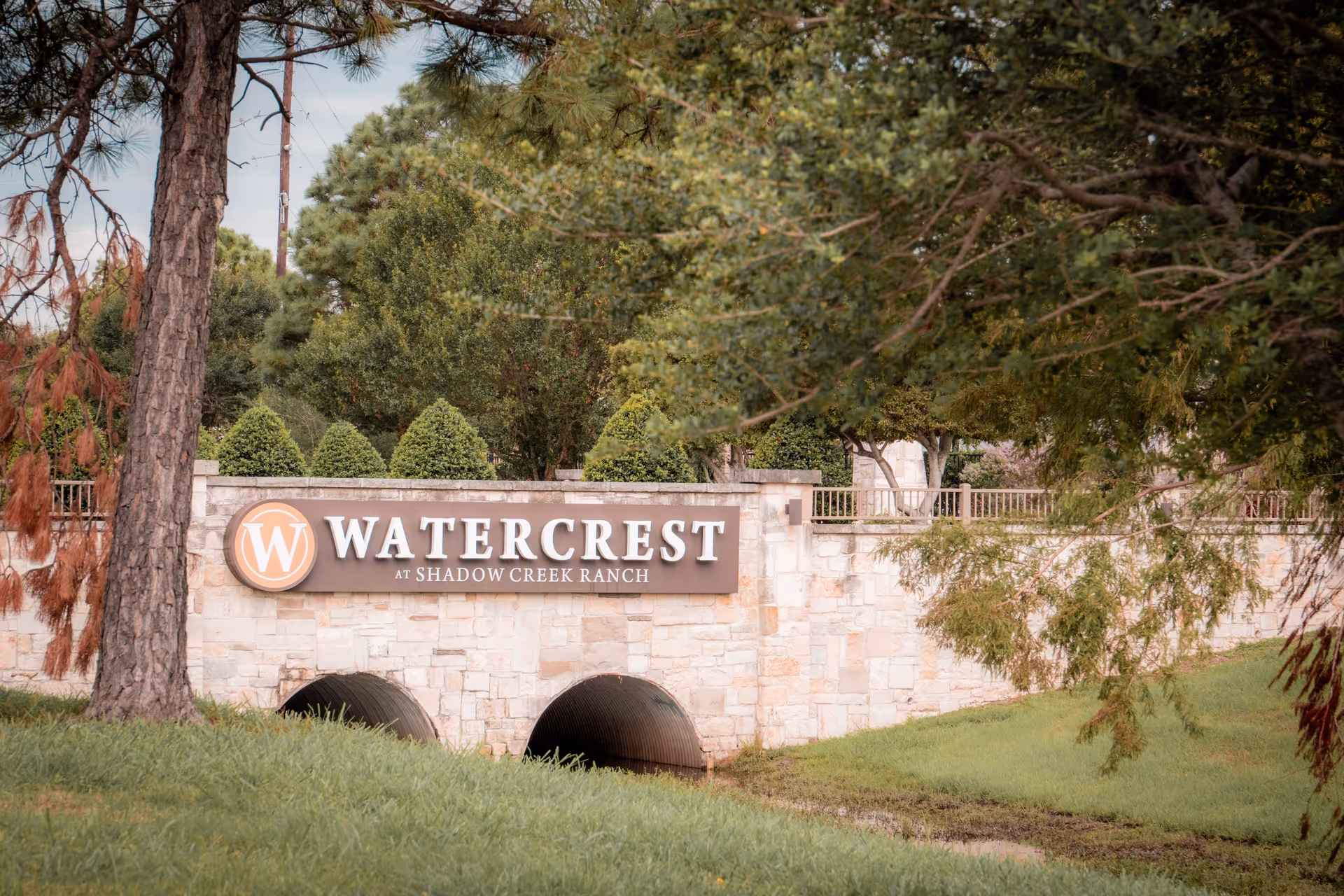 Stone entrance sign for Watercrest at Shadow Creek Ranch surrounded by green trees and grass, with two culvert tunnels underneath the sign.