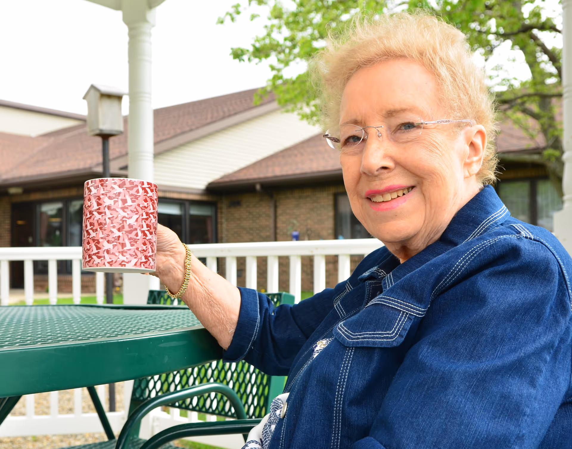 An elderly woman with short blonde hair and glasses is sitting outdoors at a green metal table, holding a red patterned mug and smiling. Behind her is a white fence, a birdhouse, and a building with a brown roof and brick walls, with trees in the background.