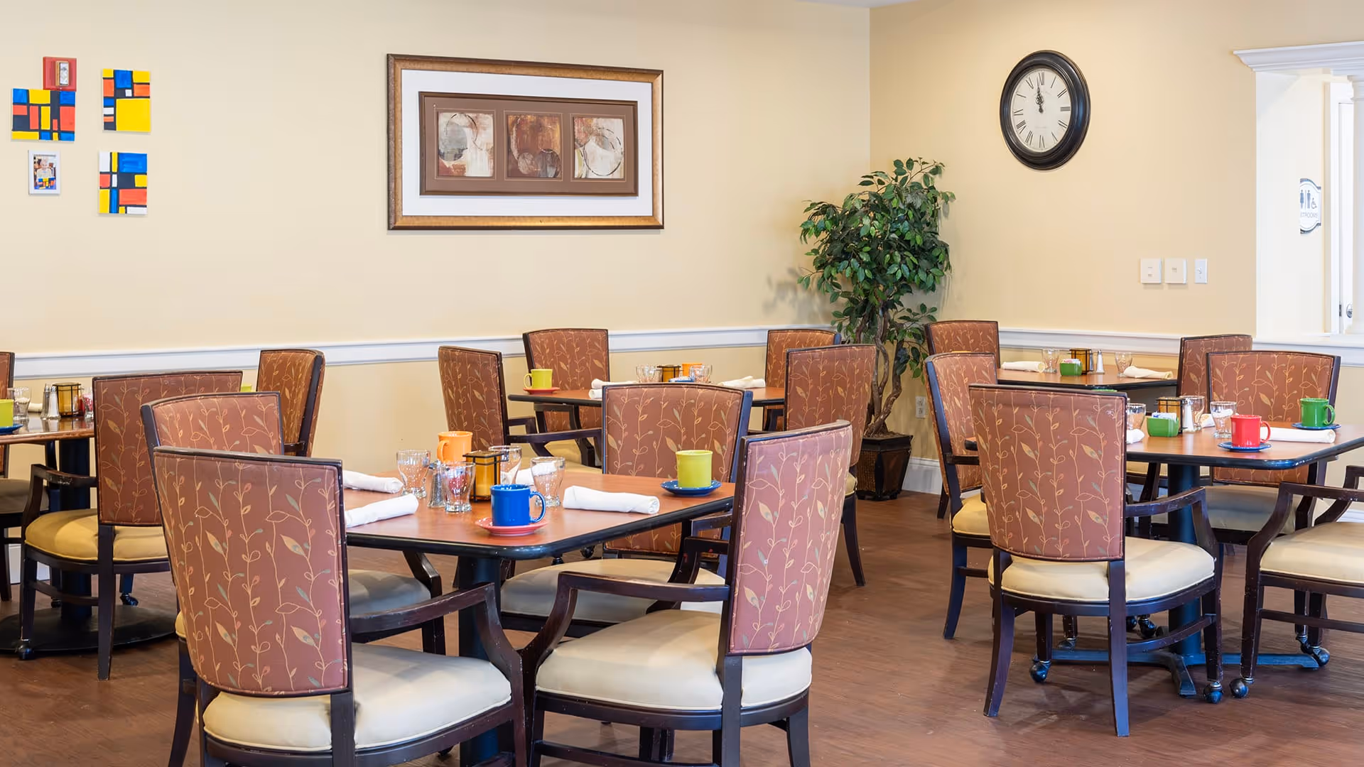 A dining room with several wooden tables and cushioned chairs arranged neatly. Each table has colorful coffee mugs, glasses, and white napkins. The walls are light yellow with framed artwork and a clock. A potted plant is placed in the corner near an archway.