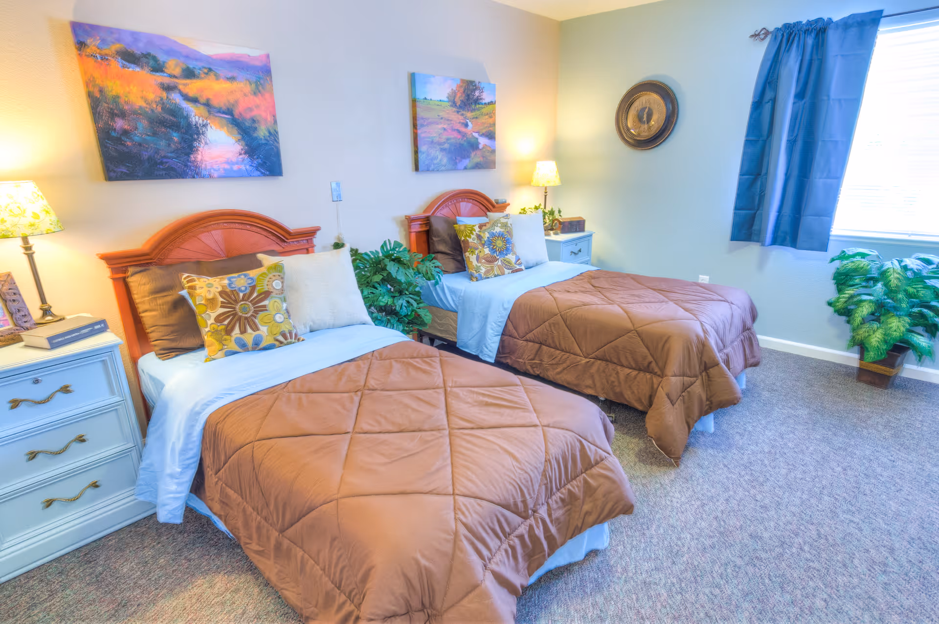 A bright bedroom with two twin beds featuring brown comforters and decorative pillows. Each bed has a wooden headboard and is flanked by white nightstands with lamps. The room has carpeted flooring, two colorful landscape paintings on the wall, a window with blue curtains, and green potted plants near the window and between the beds.