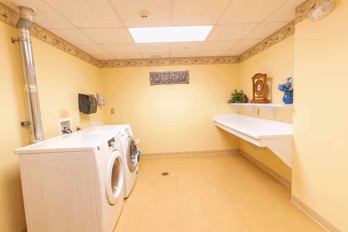 A laundry room with yellow walls and a floral border near the ceiling. There are two white front-loading washing machines on the left side. On the right side, there is a long white folding counter with a shelf above it holding a clock, a plant, and a blue vase with flowers. A sign on the back wall reads 'LAUNDRY good clean fun'. The room has a beige floor and a ceiling light panel.