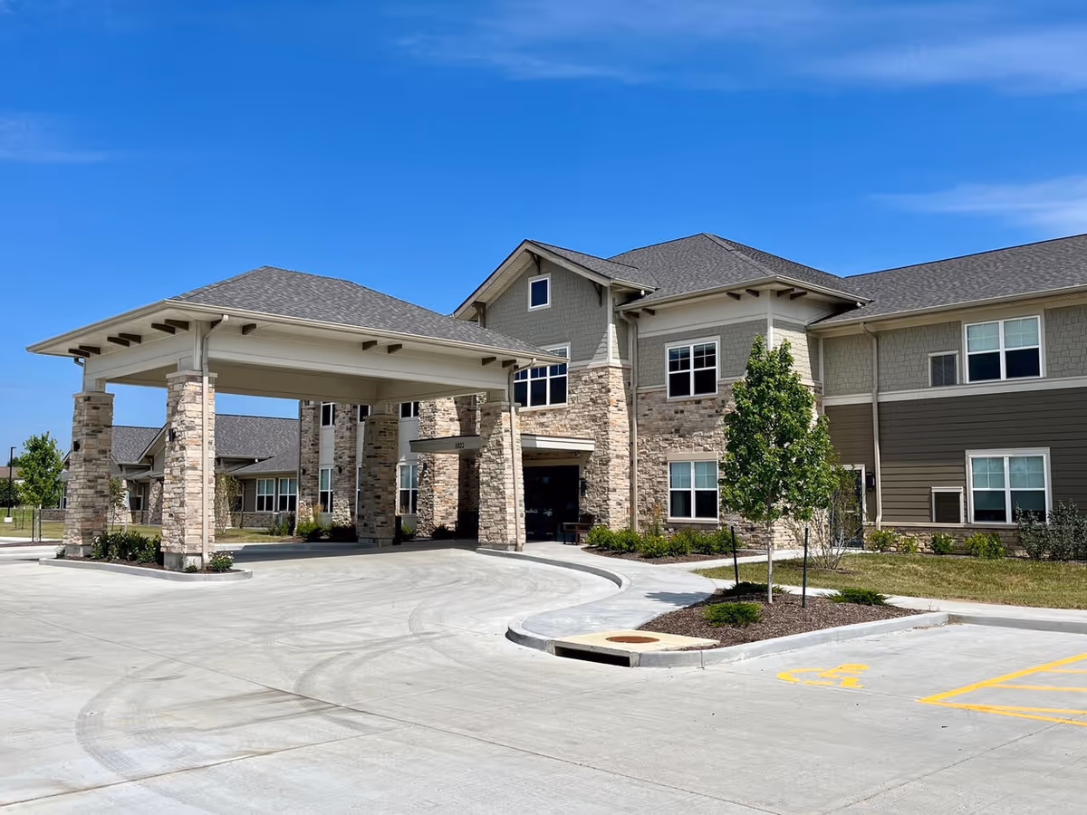 Exterior view of a senior living facility building with stone and siding facade, a covered entrance supported by stone pillars, a driveway, and a small landscaped area with a tree under a clear blue sky.