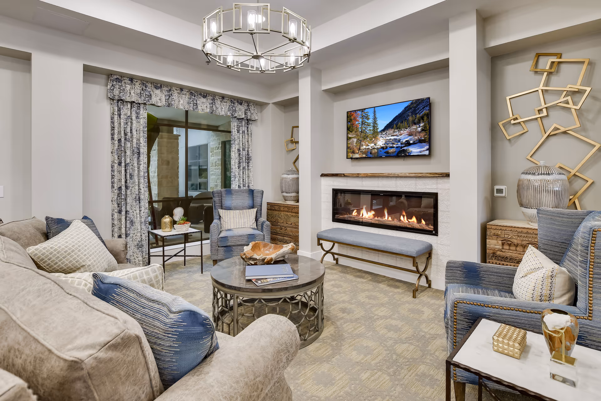 A cozy living room with a beige sofa and blue armchairs arranged around a round coffee table. A modern fireplace with a bench underneath is built into the wall, above which a flat-screen TV displays a scenic mountain and river view. The room features patterned curtains, decorative wall art, and a chandelier hanging from the ceiling.