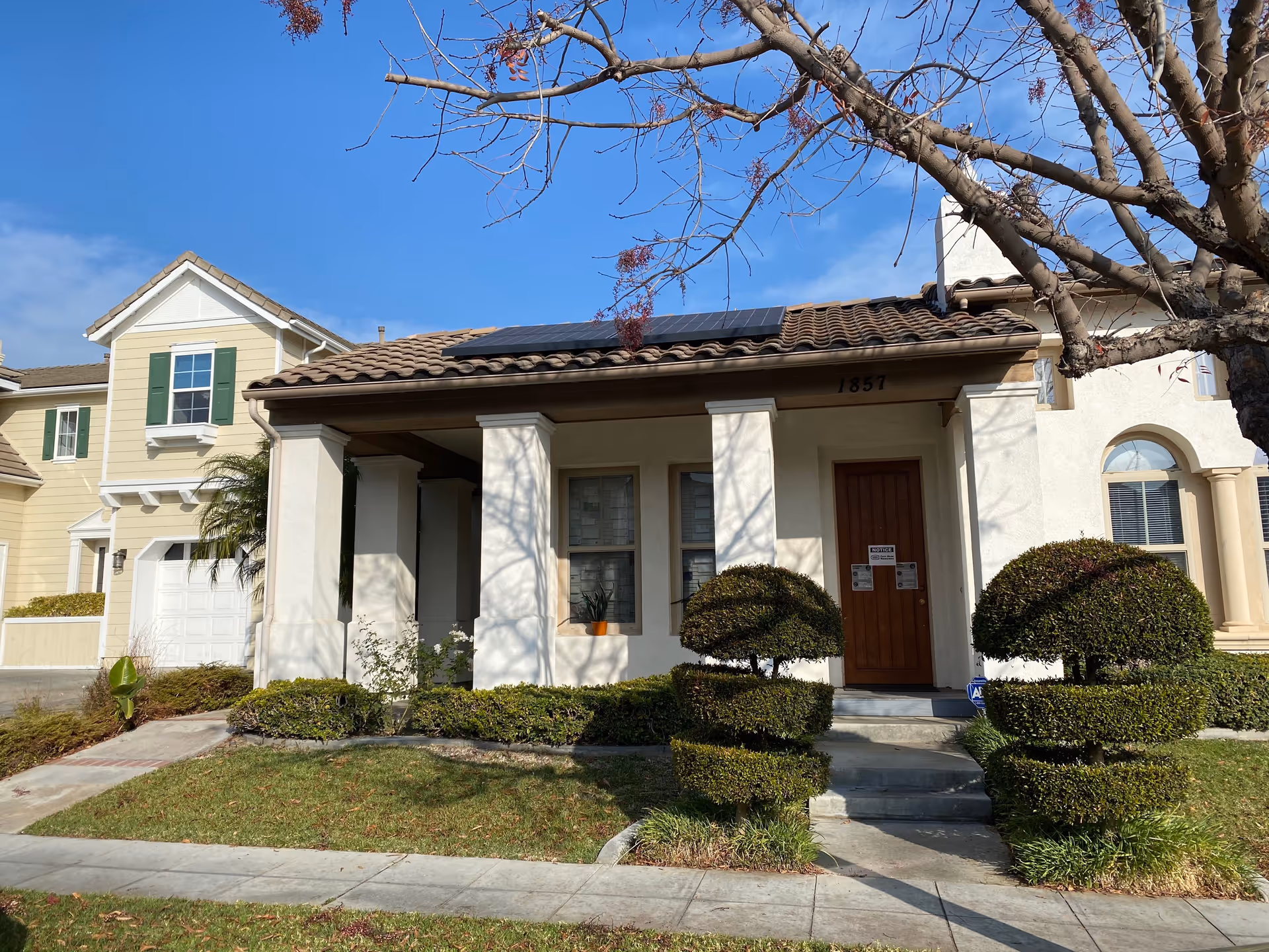 Front exterior view of a residential-style building with beige walls, a brown tiled roof with solar panels, a wooden front door, and neatly trimmed bushes and trees in the front yard under a clear blue sky.