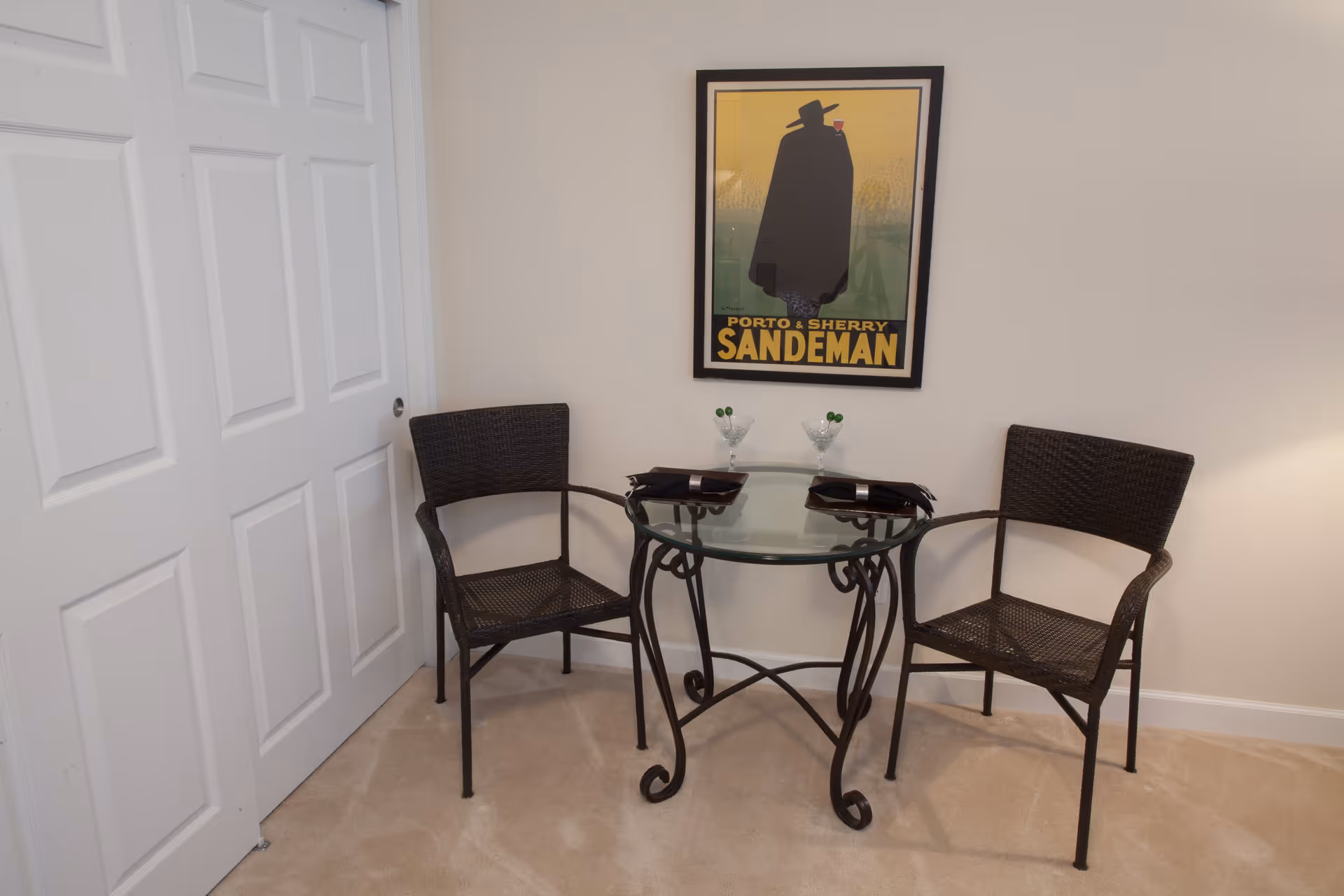 A small dining area with a round glass table and two black wicker chairs. On the table are two martini glasses with green olives and two black napkins. A framed vintage-style poster with the text 'Porto & Sherry Sandeman' hangs on the beige wall behind the table. To the left, there is a white sliding door closet.