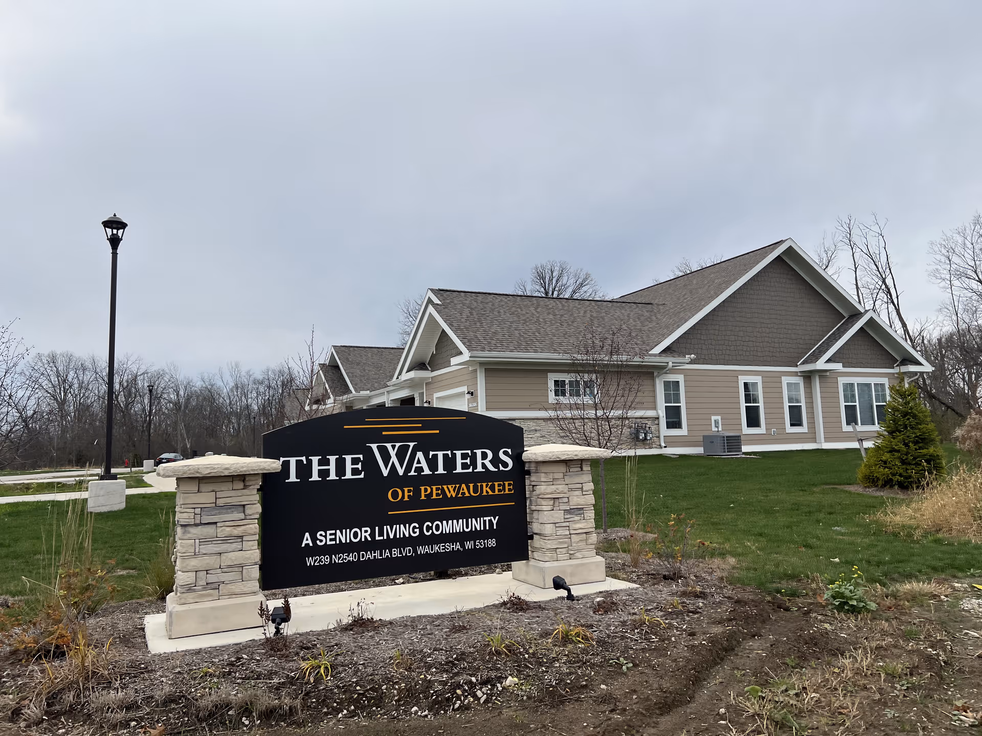 Outdoor view of The Waters of Pewaukee senior living community sign with a beige and brown building in the background, surrounded by grass and some trees under a cloudy sky.