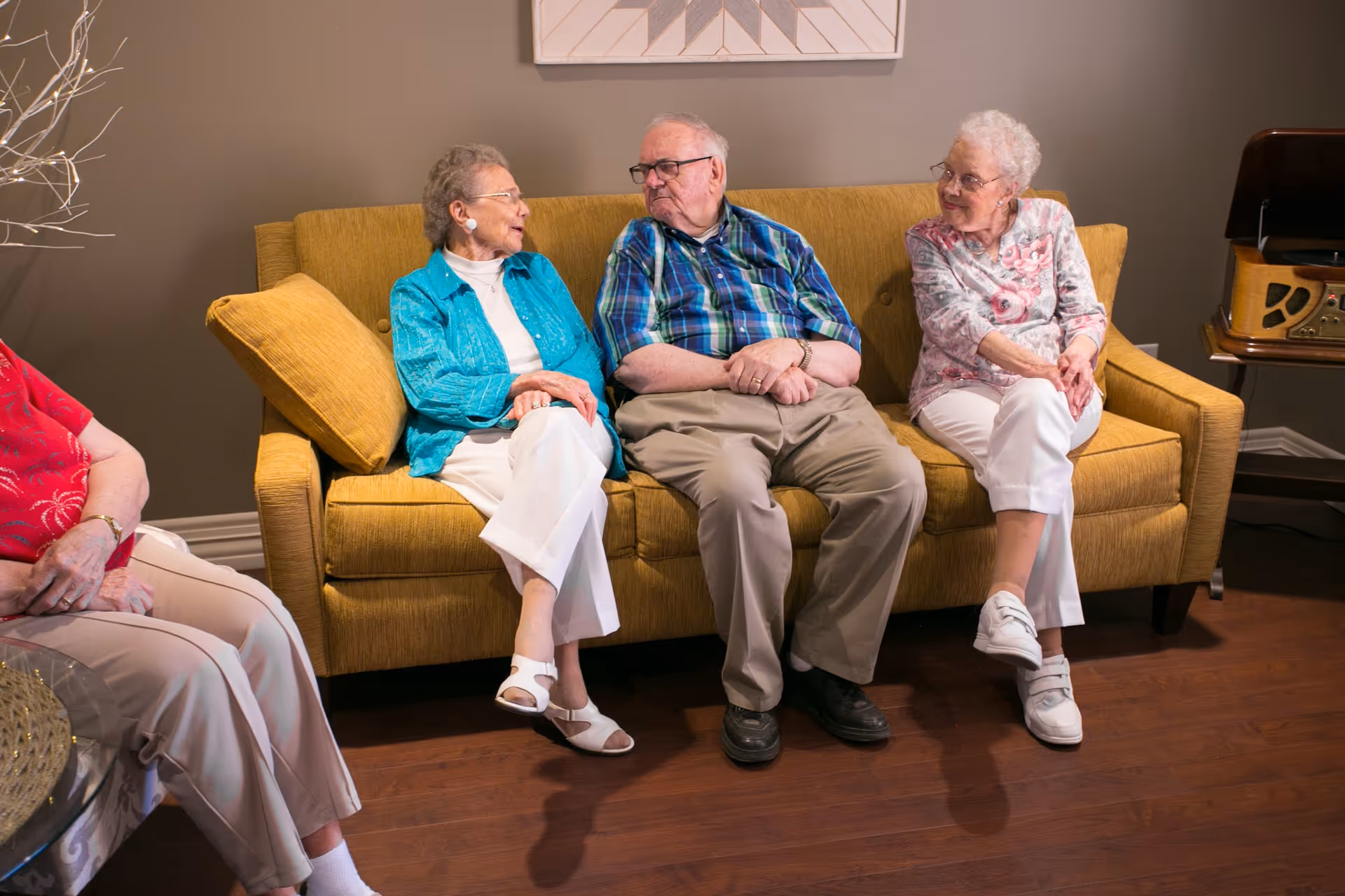 Three elderly individuals sitting on a mustard yellow couch in a living room setting, engaged in conversation. The room has wooden flooring, a brown wall, and a vintage-style record player on a small table to the right.