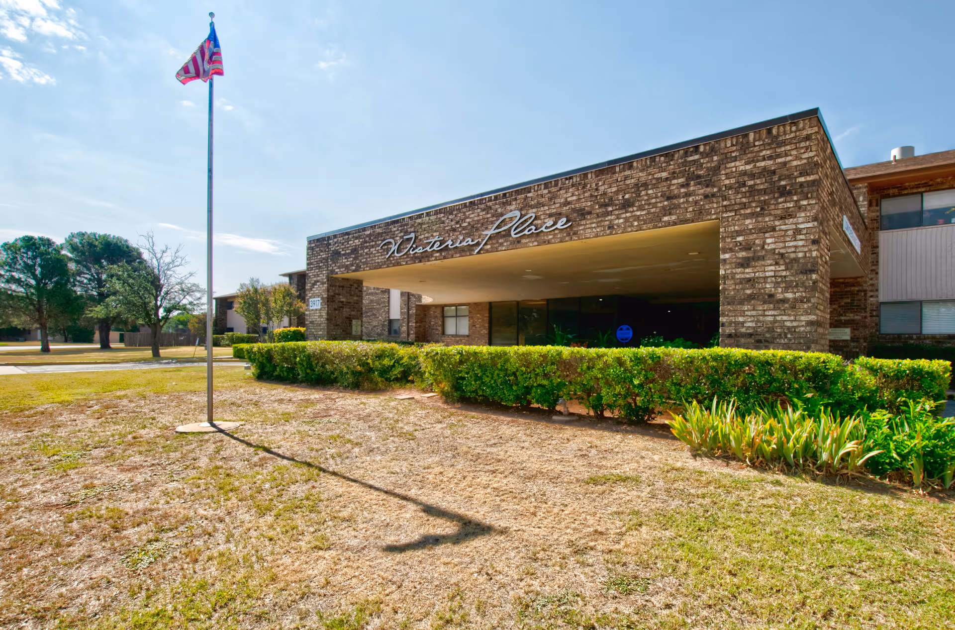 Exterior view of Wisteria Place Independent Living facility showing a brick building with a covered entrance, a flagpole with an American flag, green bushes, and trees under a clear sky.