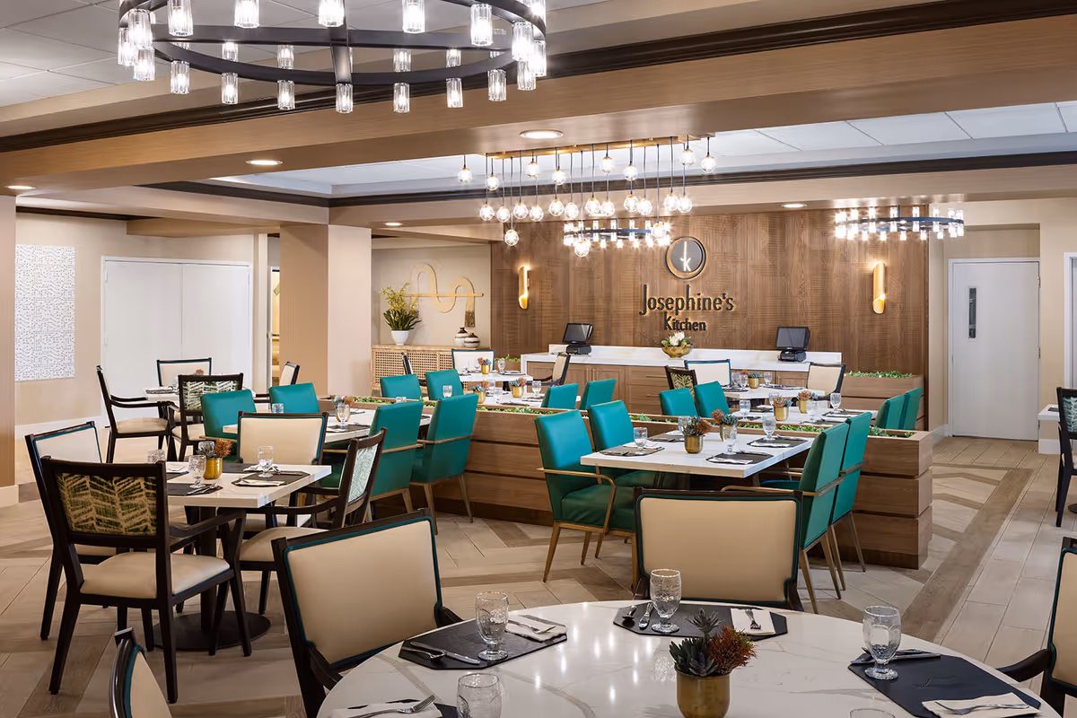 A modern dining room with multiple tables set with glasses, napkins, and cutlery. The chairs are a mix of teal and beige upholstery. The back wall features a wooden panel with the sign 'Josephine's Kitchen' and two computer stations. The room is well-lit with contemporary chandeliers and wall sconces.