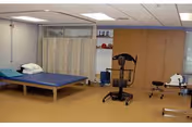A rehabilitation room with a blue therapy table, a privacy curtain, exercise equipment, and a stool. The room has beige flooring and light-colored walls with a window on the right side.