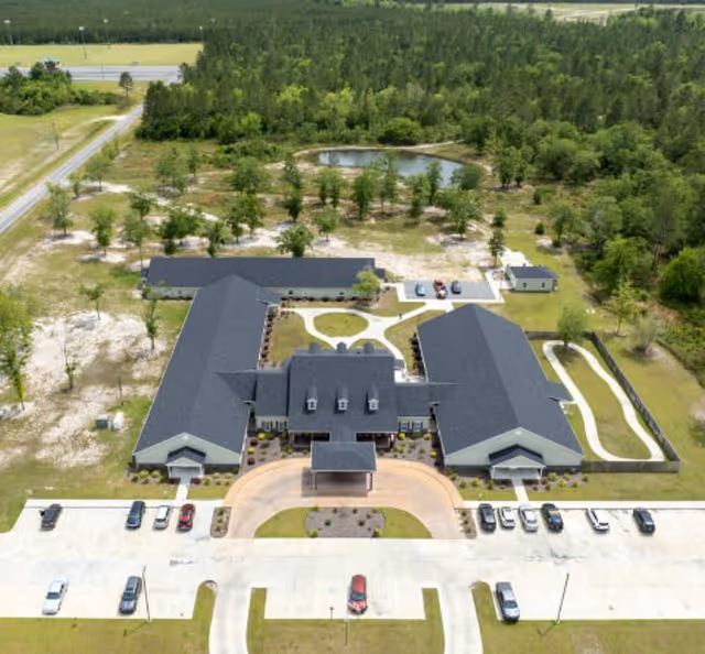 Aerial view of At Home Senior Living of Jesup facility showing a large building with a dark roof surrounded by greenery and trees. The building has a circular driveway with several parked cars and a small pond behind the building. There are walking paths and landscaped areas around the property.