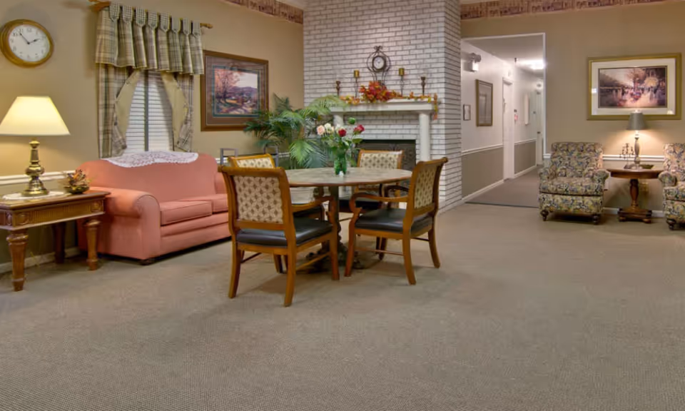 A cozy living room area with a pink sofa, a wooden side table with a lamp, a round table with four chairs, a white brick fireplace decorated with autumn leaves and candles, two floral armchairs with a small table and lamp between them, framed paintings on the walls, and a clock above the window with plaid curtains.