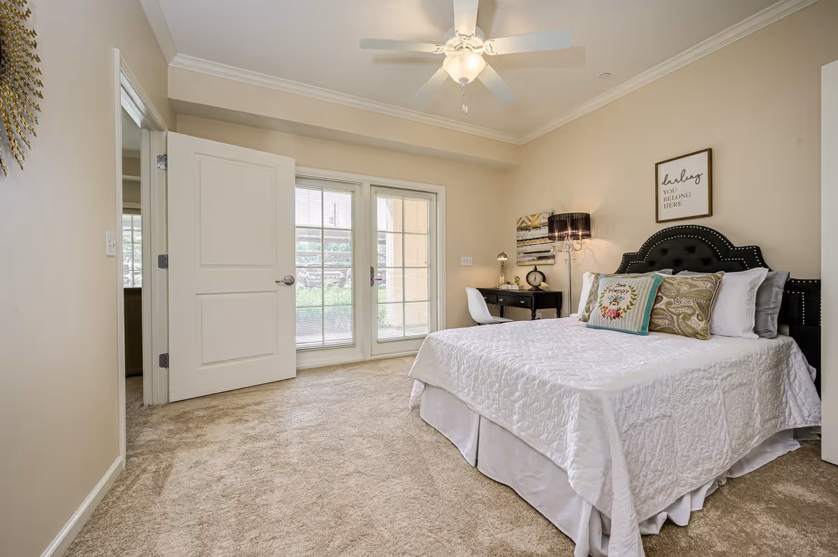 A bright bedroom with beige walls and carpet, featuring a neatly made bed with white bedding and decorative pillows. Above the bed is a framed sign that reads 'darling YOU BELONG HERE'. To the right of the bed is a black desk with a white chair, a lamp, and decorative items. French doors with blinds lead outside, and a ceiling fan with a light fixture is mounted on the ceiling.