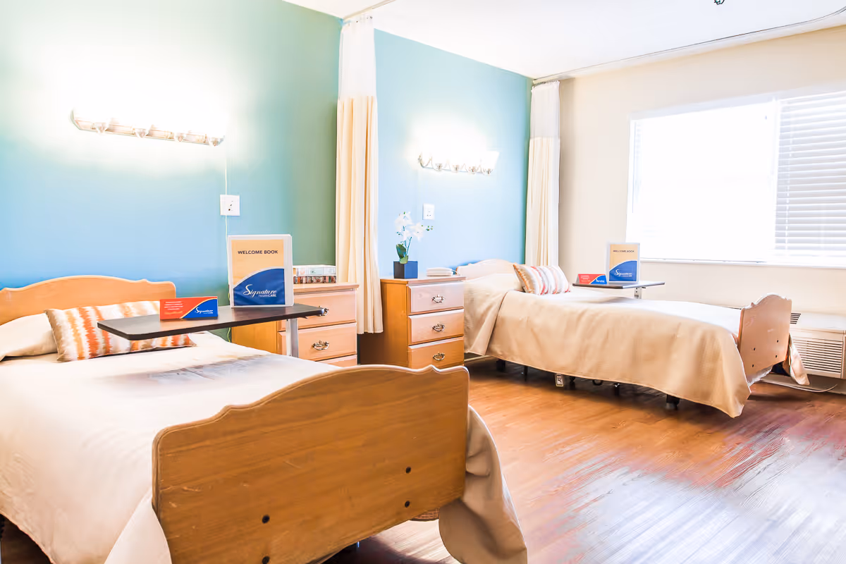 A bright and clean room with two single beds, each with a beige bedspread and a pillow. Between the beds are two wooden dressers with drawers, one with a small potted plant and the other with a welcome book and a blue card. The walls are painted light blue and beige, with wall-mounted lights above each bed. A large window with blinds allows natural light to fill the room, and the floor is wooden.