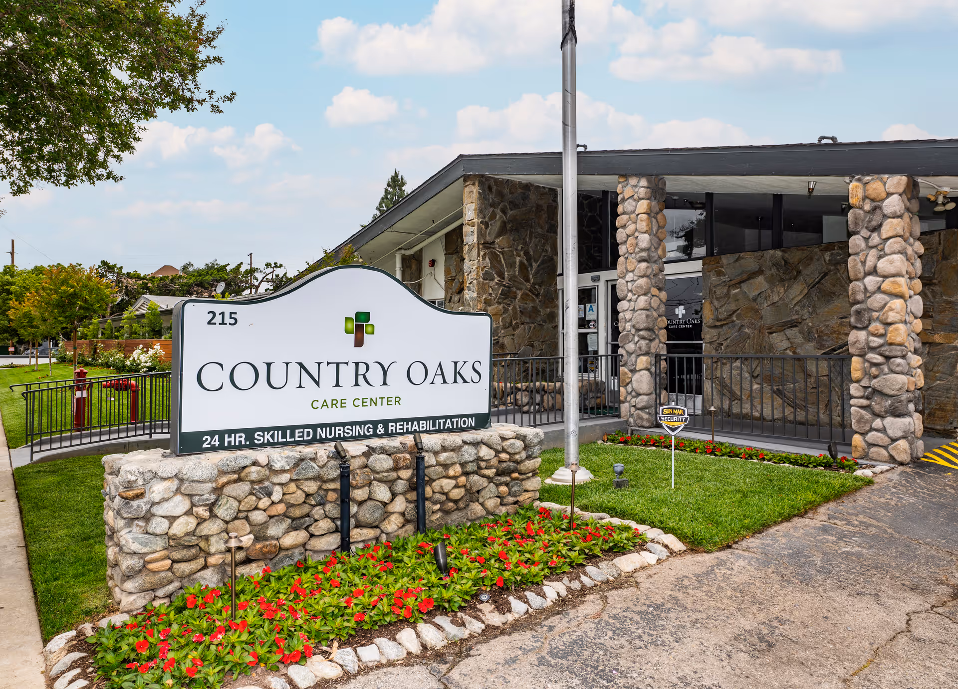 Exterior view of Country Oaks Care Center showing the entrance with stone pillars and a large sign that reads 'Country Oaks Care Center 24 HR. Skilled Nursing & Rehabilitation' surrounded by a flower bed with red flowers and green grass.