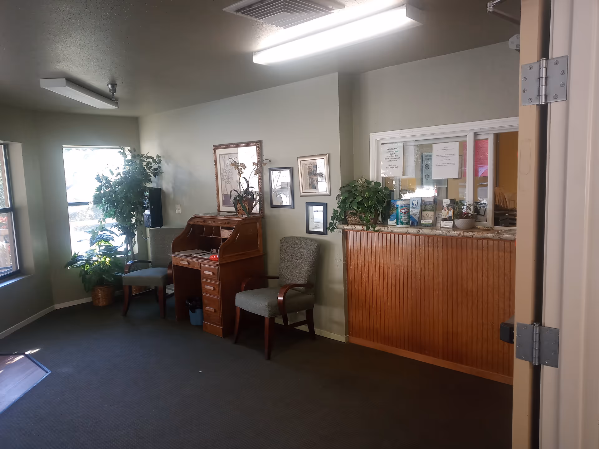 Interior view of a senior living facility reception area with two upholstered chairs, a wooden roll-top desk, potted plants, framed artwork on the wall, and a reception window with notices and cleaning supplies on the counter.