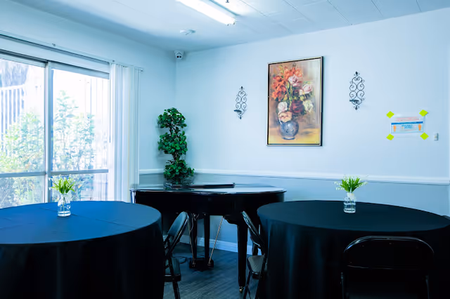 A bright room with two round tables covered with black tablecloths, each with a small vase of flowers. There are black chairs around the tables. In the corner, there is a black grand piano with a tall green plant beside it. On the wall above the piano, there is a framed painting of flowers and two decorative wall sconces. A large window with vertical blinds lets in natural light.