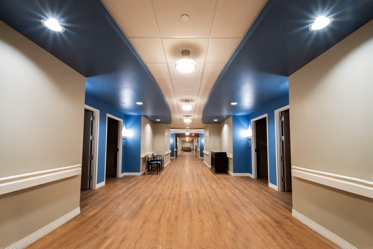Wide hallway in a senior living facility with wood flooring, beige walls, blue ceiling accents, multiple open doorways on both sides, ceiling lights, and some chairs and furniture along the walls.