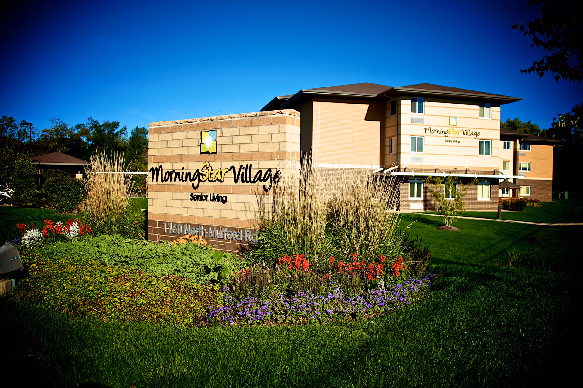 Exterior view of Morning Star Village senior living facility with a large sign in the foreground surrounded by colorful flowers and greenery. The multi-story building is visible in the background under a clear blue sky.