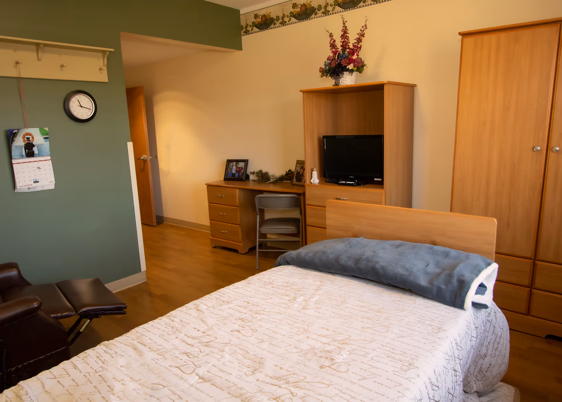 A cozy bedroom in a senior living facility featuring a single bed with a patterned bedspread and a folded blanket at the foot. There is a wooden wardrobe and a matching dresser with a small TV on top. A desk with a chair and framed photos is positioned against the wall. A recliner chair is visible in the corner, and a wall clock and calendar hang on a green accent wall.