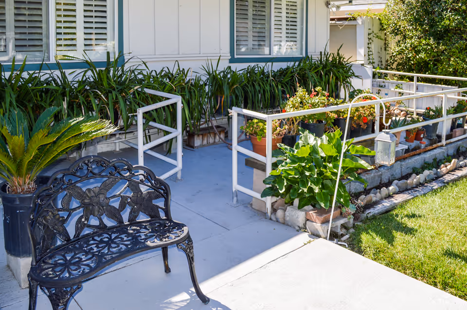 Outdoor patio area with a decorative black metal bench, potted plants, and a white railing. The area is adjacent to a building with white walls and windows with shutters. There is a small garden with green grass and various plants in pots along the railing.