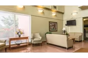 A bright living room area with large windows letting in natural light. The room features a beige sofa, two armchairs, a wooden table with a vase of flowers, a wall-mounted TV, and framed artwork on the wall. The walls are painted light green and the floor has a polished wood finish.