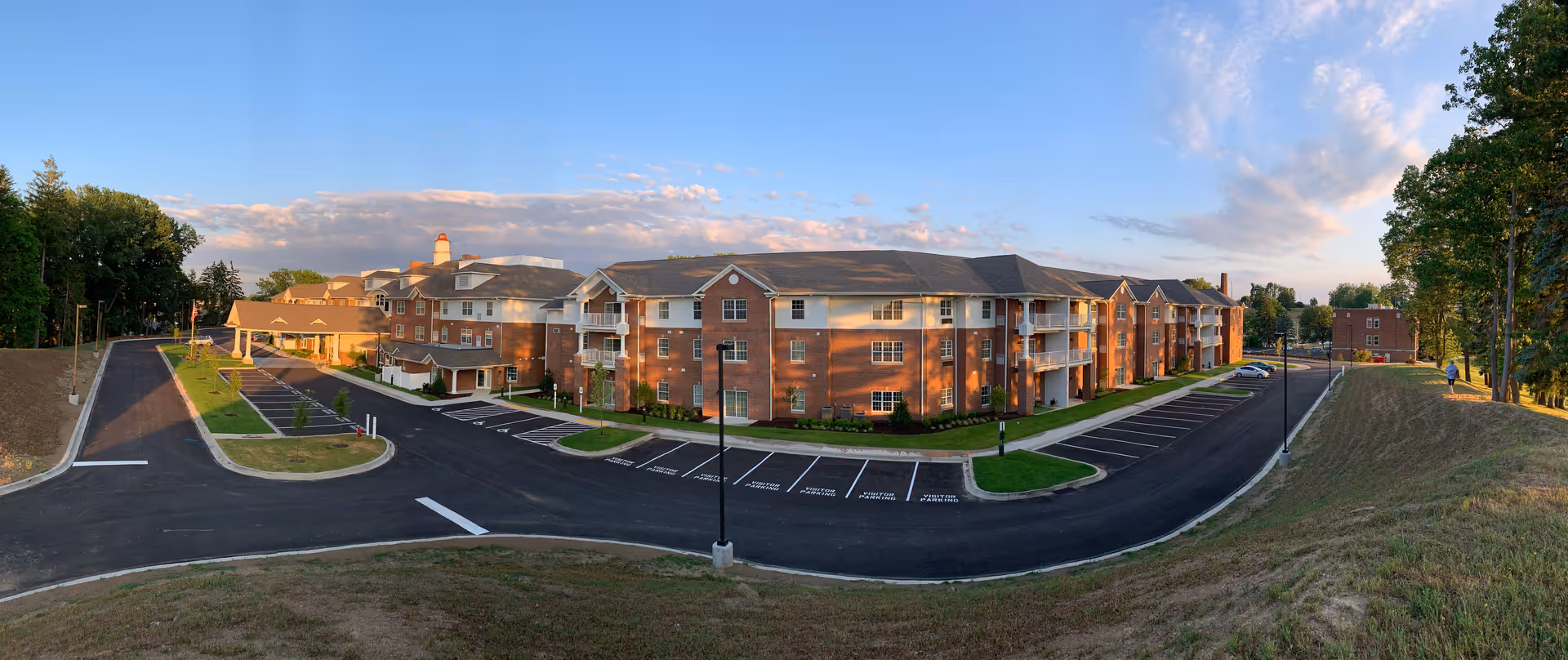 Panoramic view of South Hills Square Retirement Resort showing a large, multi-story brick building with white balconies and a parking lot in front. The building is surrounded by trees and grassy areas under a partly cloudy sky during sunset.