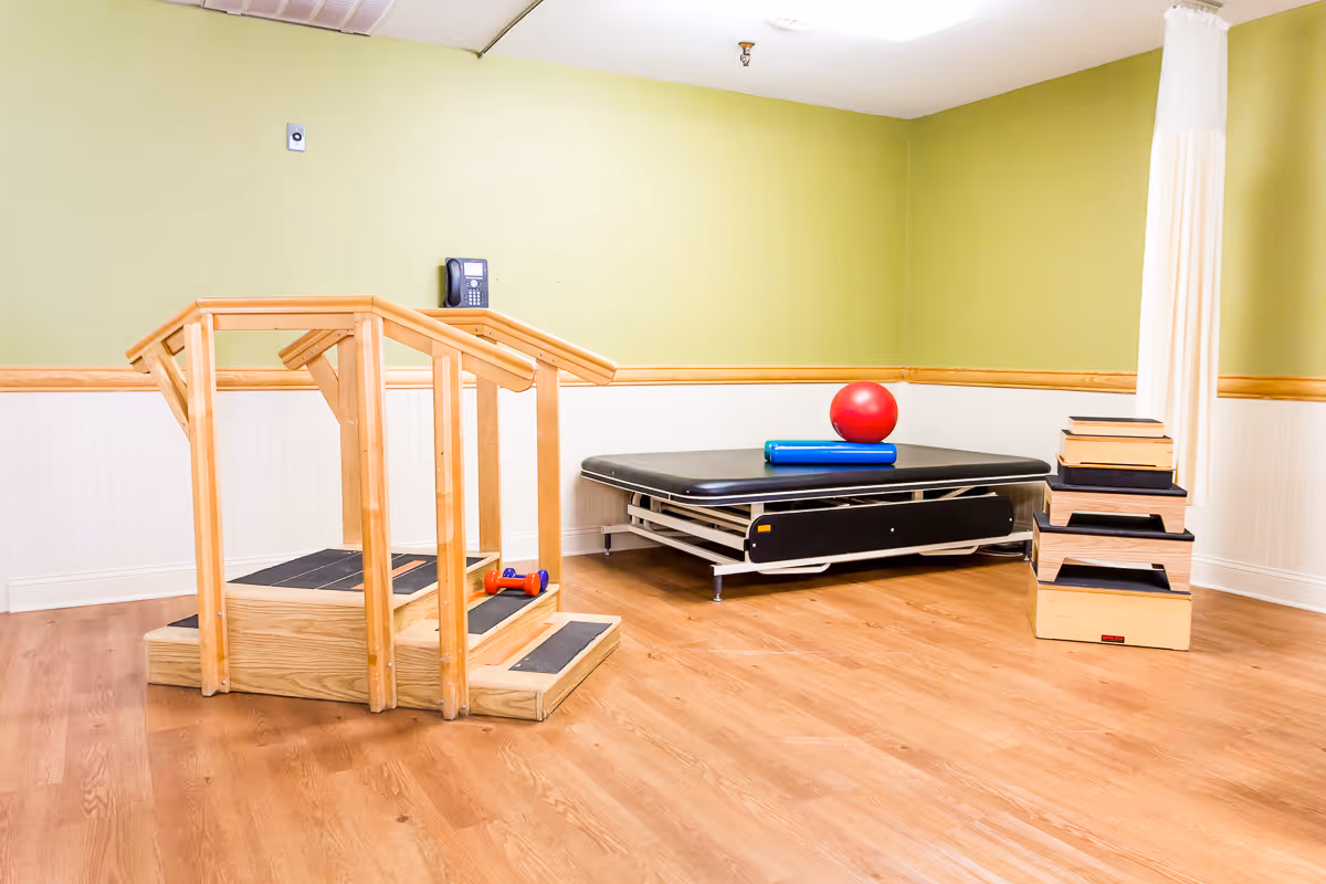 A physical therapy room with wooden stairs for rehabilitation, a black therapy table with a red exercise ball and blue foam roller on top, and a set of wooden step platforms stacked in the corner. The walls are painted green and white with wood trim, and the floor is light wood.