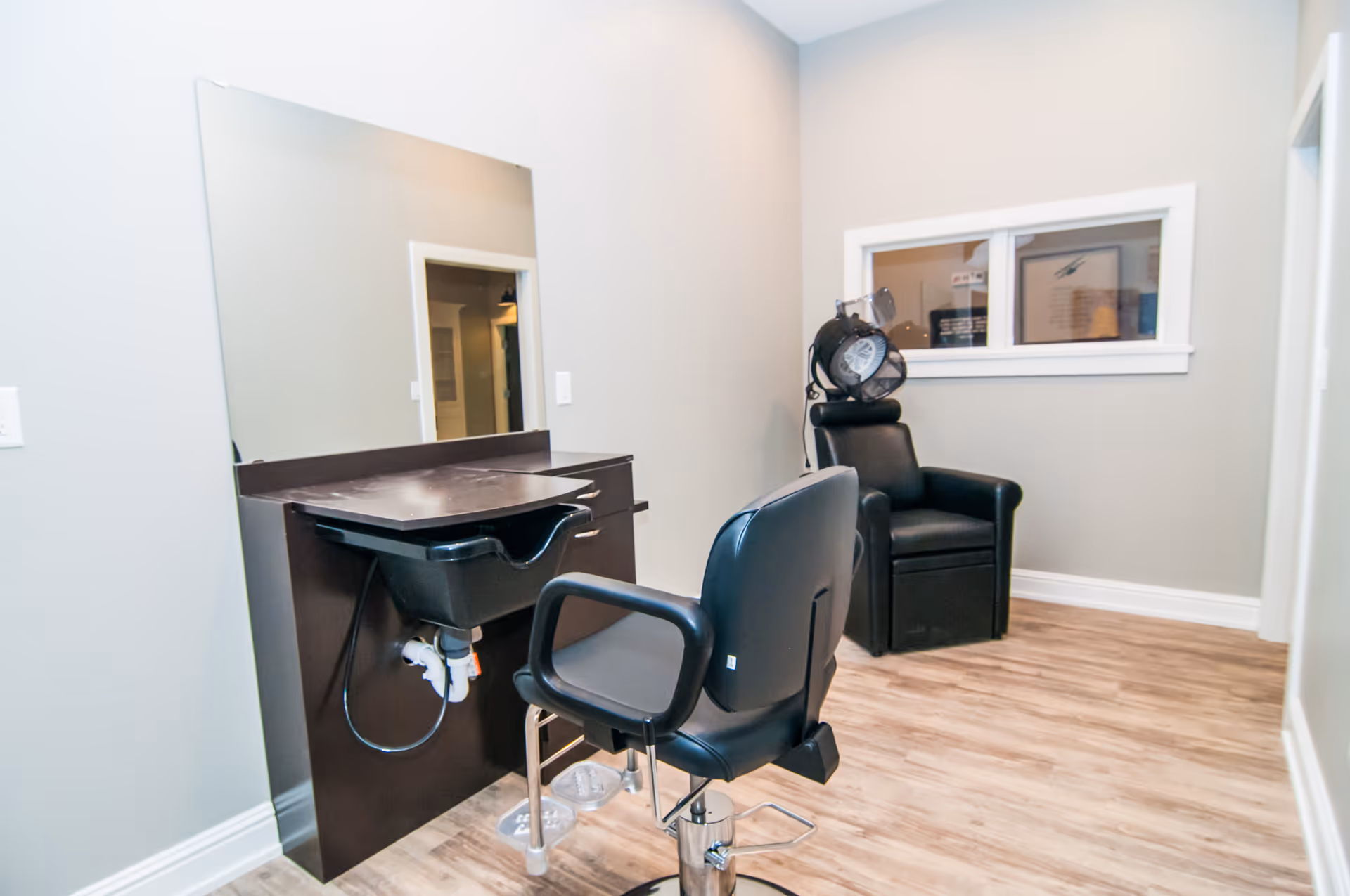 A small salon area inside a senior living facility with a black salon chair in front of a wash basin and a large mirror on the wall. In the background, there is a black leather chair with a hair dryer attached. The room has light-colored walls and wood flooring.