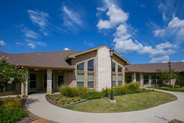 Exterior view of a single-story healthcare and rehabilitation facility building with a stone and brick facade, large windows, a well-maintained lawn, shrubs, and a curved concrete walkway under a partly cloudy blue sky.
