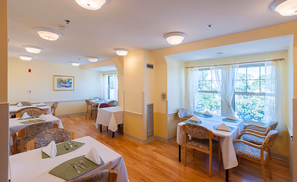 A bright dining room with several tables covered in white tablecloths and green placemats, each set with folded white napkins and silverware. The room has wooden floors, light yellow walls, multiple ceiling lights, and large windows with sheer curtains letting in natural light. Comfortable upholstered chairs surround the tables.