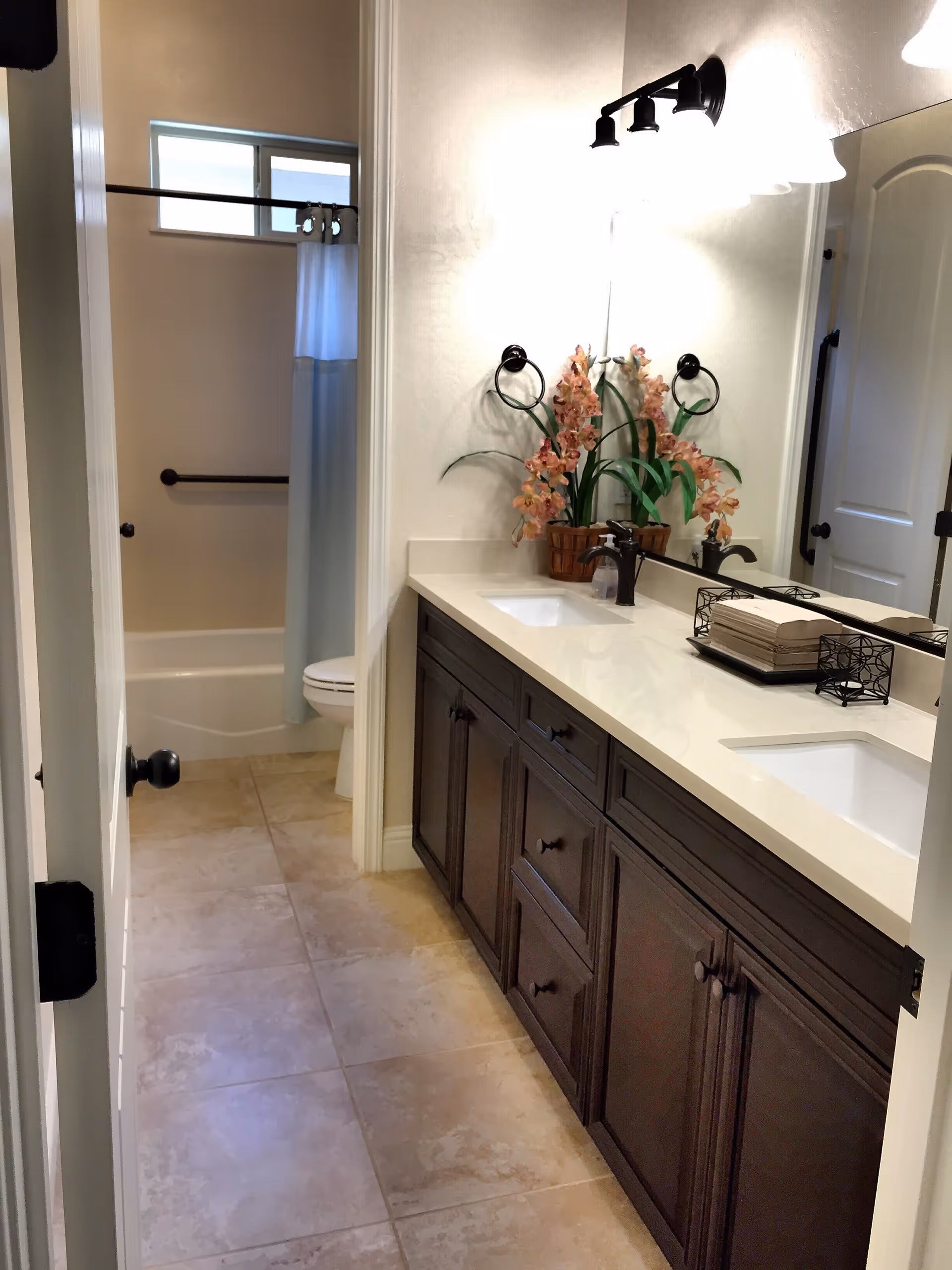 A bathroom with a double sink vanity featuring dark wood cabinets and a white countertop. There are two black faucets, a decorative plant with pink flowers, and a tray with folded towels on the countertop. The bathroom has beige tiled flooring and a separate area with a toilet and a bathtub with a white shower curtain. The walls are light-colored, and there is a large mirror above the sinks with two wall-mounted light fixtures.