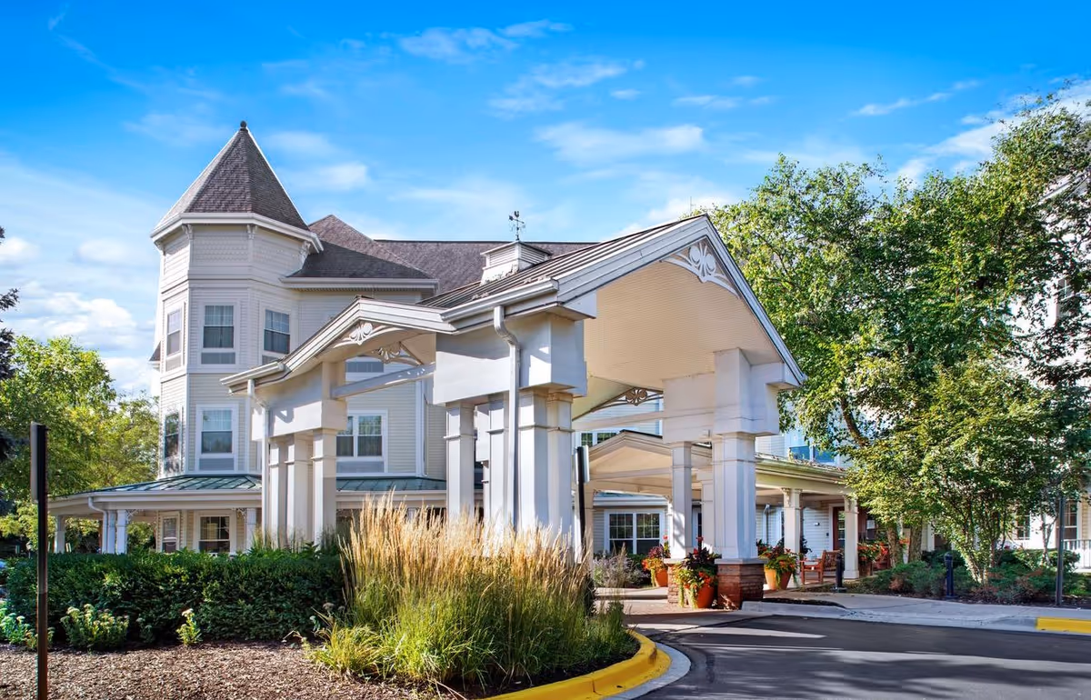 Exterior view of a senior living facility named Sunrise of Schaumburg featuring a large covered entrance with white pillars, a turret-style architectural element, surrounding greenery, and a clear blue sky.