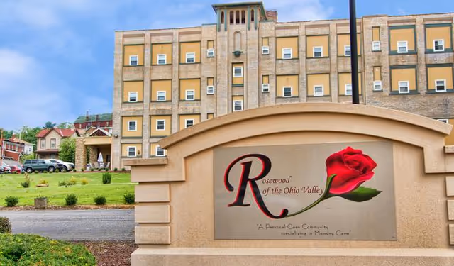 Exterior view of a multi-story senior living facility building with a large sign in the foreground that reads 'Rosewood of the Ohio Valley' with a red rose graphic and the tagline 'A Personal Care Community specializing in Memory Care'. The building has multiple windows and is surrounded by a well-maintained lawn and driveway.