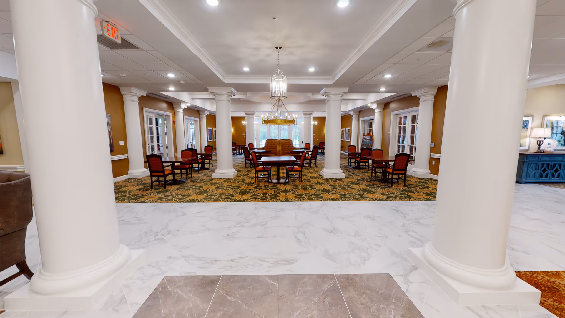 Spacious interior dining area with multiple tables and chairs arranged on a patterned carpet. The room features large white columns, warm brown walls, and elegant chandeliers hanging from a white ceiling. There are windows and glass doors along the sides, and a cushioned bench seating area in the center background.