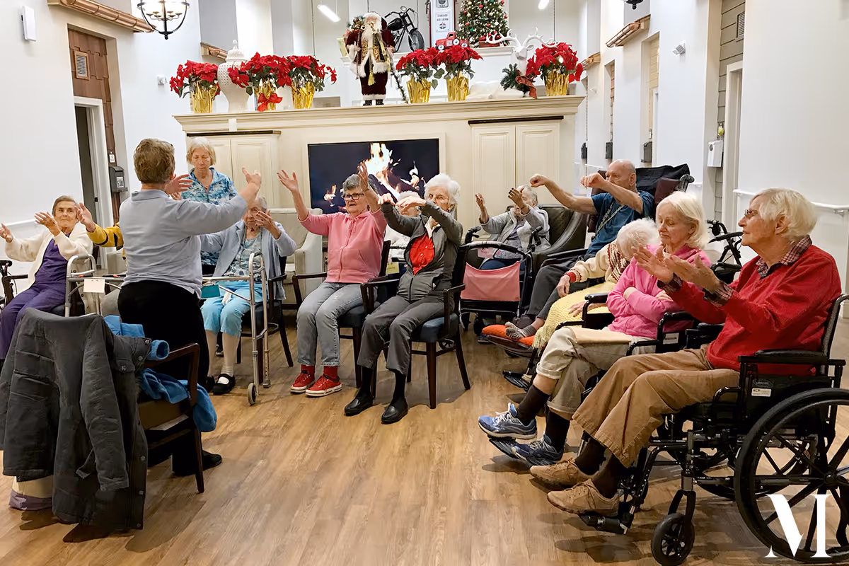 A group of elderly people seated in a semi-circle in a common room, participating in a seated exercise or stretching activity led by an instructor. The room is decorated with Christmas poinsettias, a Santa Claus figure, and a Christmas tree. A fireplace with a digital fire display is visible in the background.