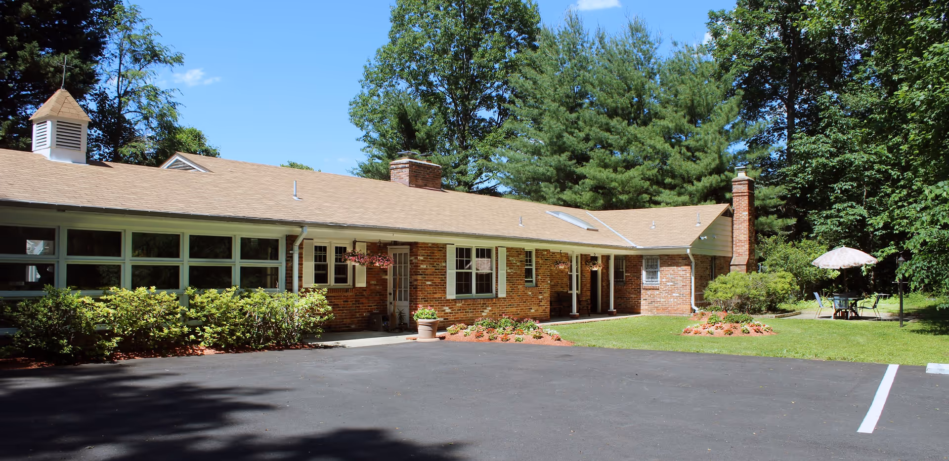 Exterior view of a single-story brick building with a beige roof, surrounded by greenery and trees. There is a paved parking area in front, flower beds near the entrance, and an outdoor seating area with a table and umbrella on the right side.