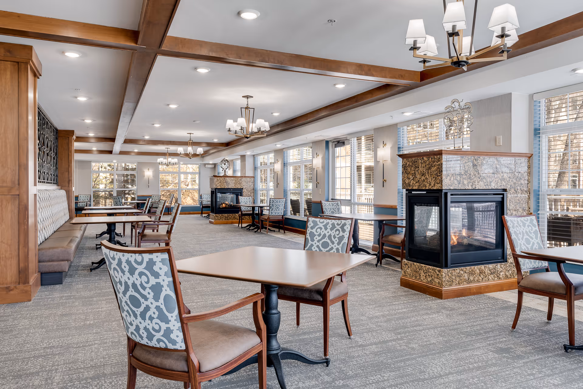 A spacious dining room with multiple tables and chairs arranged neatly. The room features large windows allowing natural light to fill the space, wooden ceiling beams, and a central fireplace with a granite surround. The decor includes patterned upholstery on the chairs and modern light fixtures hanging from the ceiling.