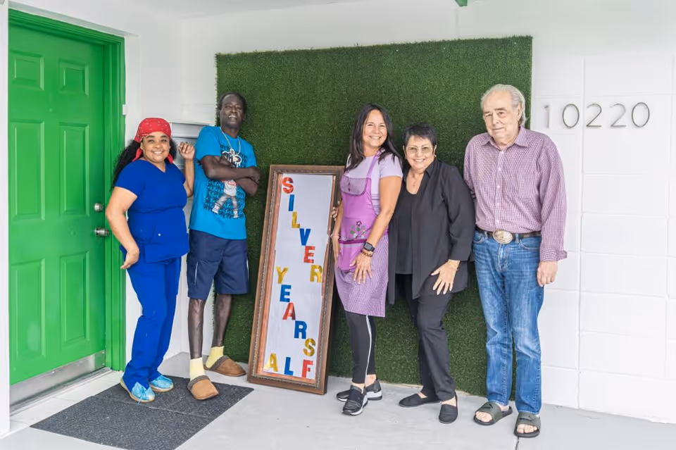 Five people standing in front of a green door and a wall with the number 10220. They are posing next to a framed sign that reads 'SILVER YEARS ALF' with colorful letters. The group includes two men and three women, smiling and casually dressed.