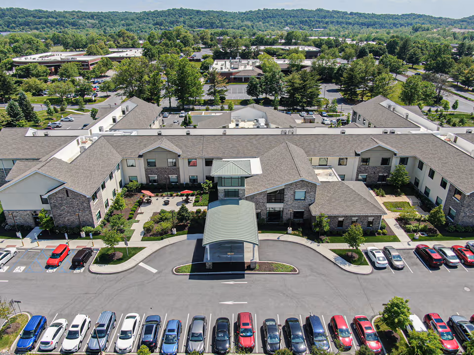 Aerial view of ProMedica Total Rehab + (Exton) facility showing a large, two-story building with a covered entrance, surrounded by landscaped gardens and parking lots filled with cars. Trees and hills are visible in the background under a clear sky.