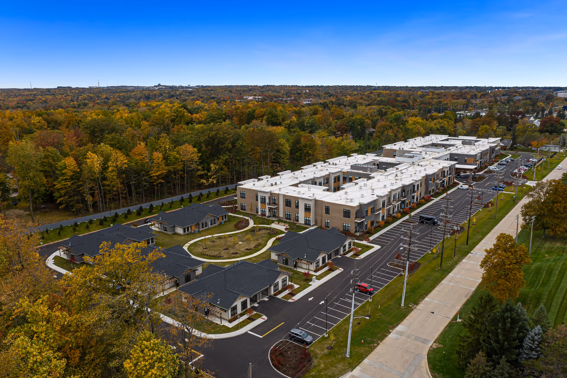Aerial view of The Ashton at Mayfield Heights senior living facility surrounded by trees with autumn foliage. The facility includes multiple buildings with white and beige exteriors, black roofs on smaller buildings, parking lots, and a circular driveway. The surrounding area features a mix of wooded land and suburban streets.