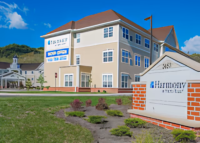 Exterior view of a multi-story senior living facility building with beige walls and a red roof under a blue sky with some clouds. In the foreground, there is a landscaped area with small bushes and a sign that reads 'Harmony at Harts Run'.